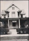 Black-and-white photo of a small two-story house with a front porch and hedges.