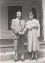 Two people stand on a porch steps, smiling in front of a house.