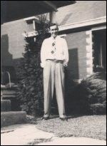 Man standing on a gravel path in front of a house, wearing a light shirt and dark trousers.