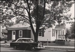 Vintage black car parked by a small house and white storefront beneath large trees