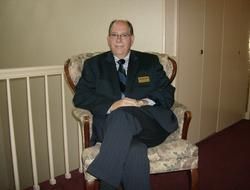 Man in a dark suit seated in an upholstered chair in a hallway, legs crossed, with a railing beside him.