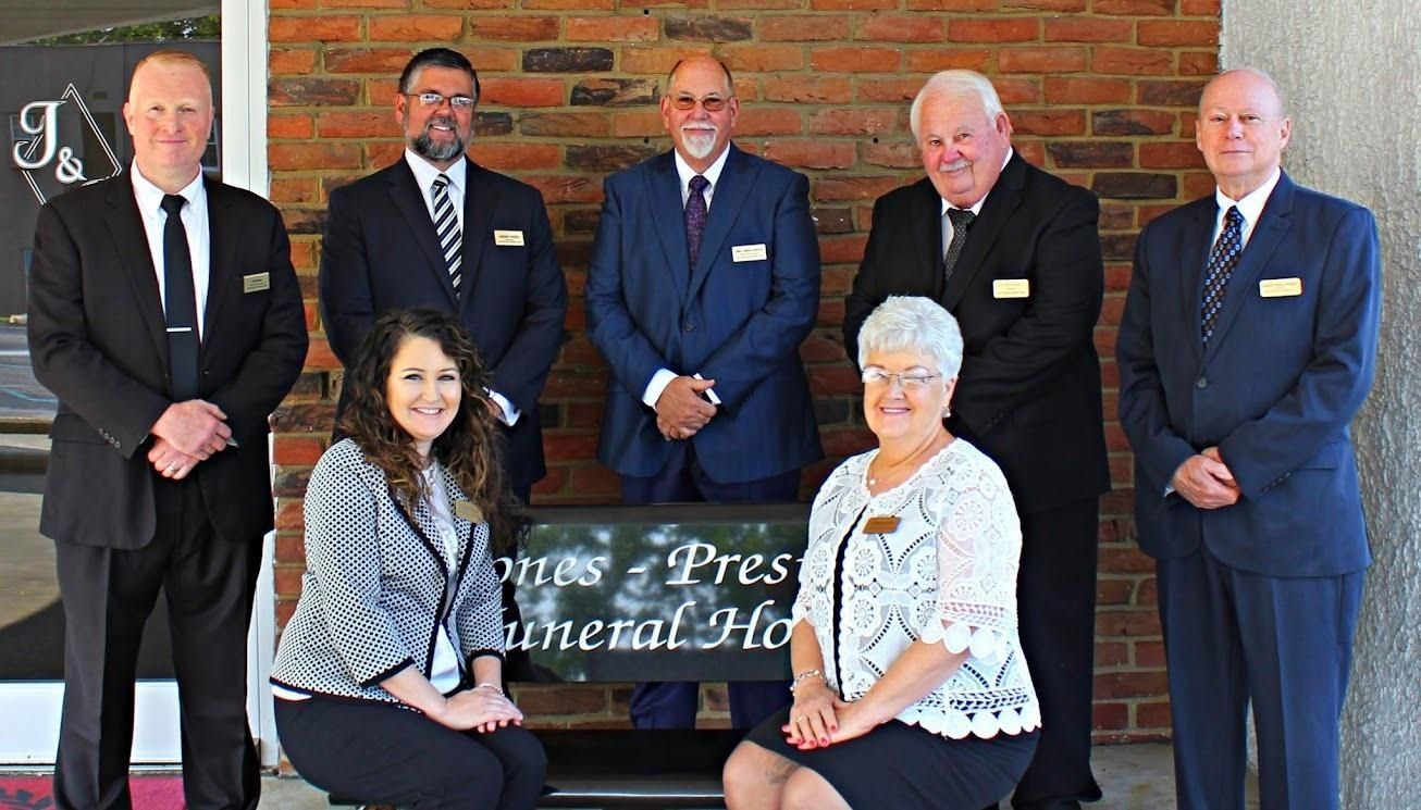 Group of seven people posing outside a brick building with a partially visible sign in front.