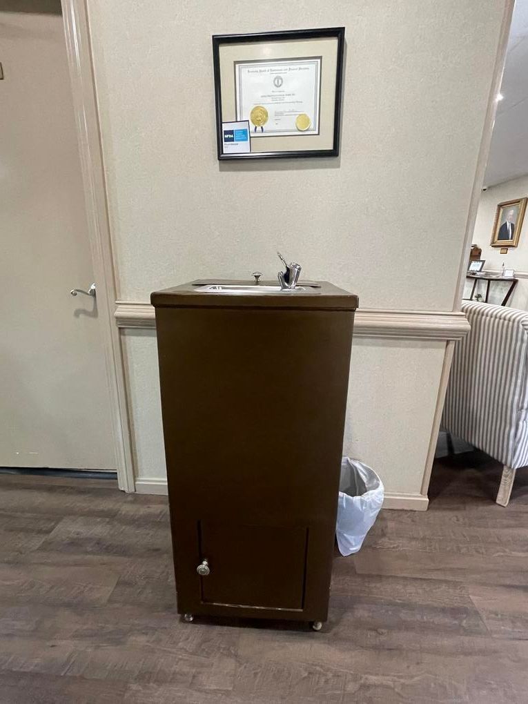 Brown cabinet with a sink in a hallway, with a framed certificate on the wall and a trash bin beside it.