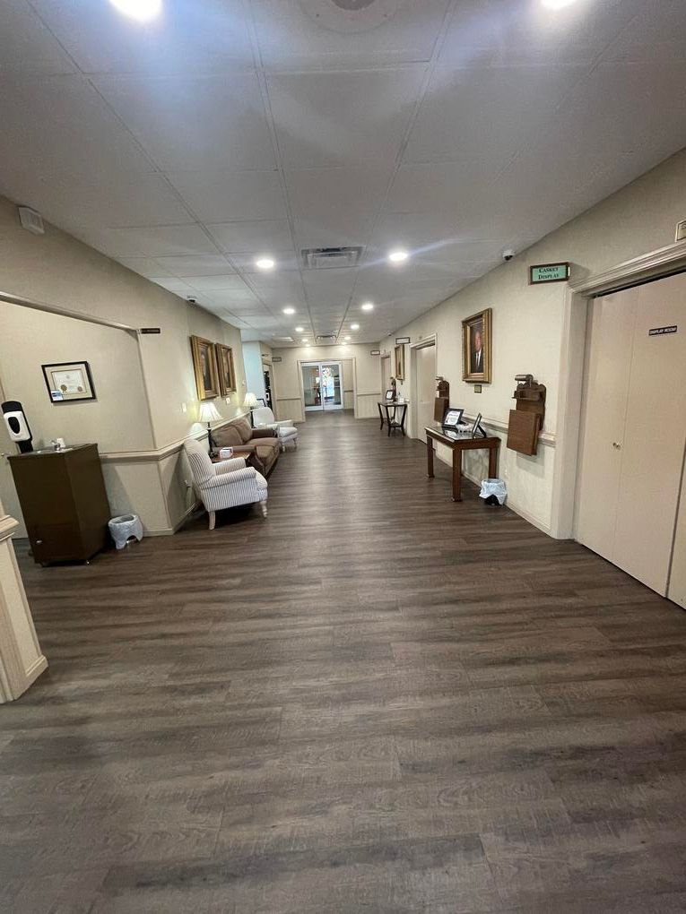 Long hallway with wood flooring, beige walls, ceiling lights, chairs, and framed art in a care facility