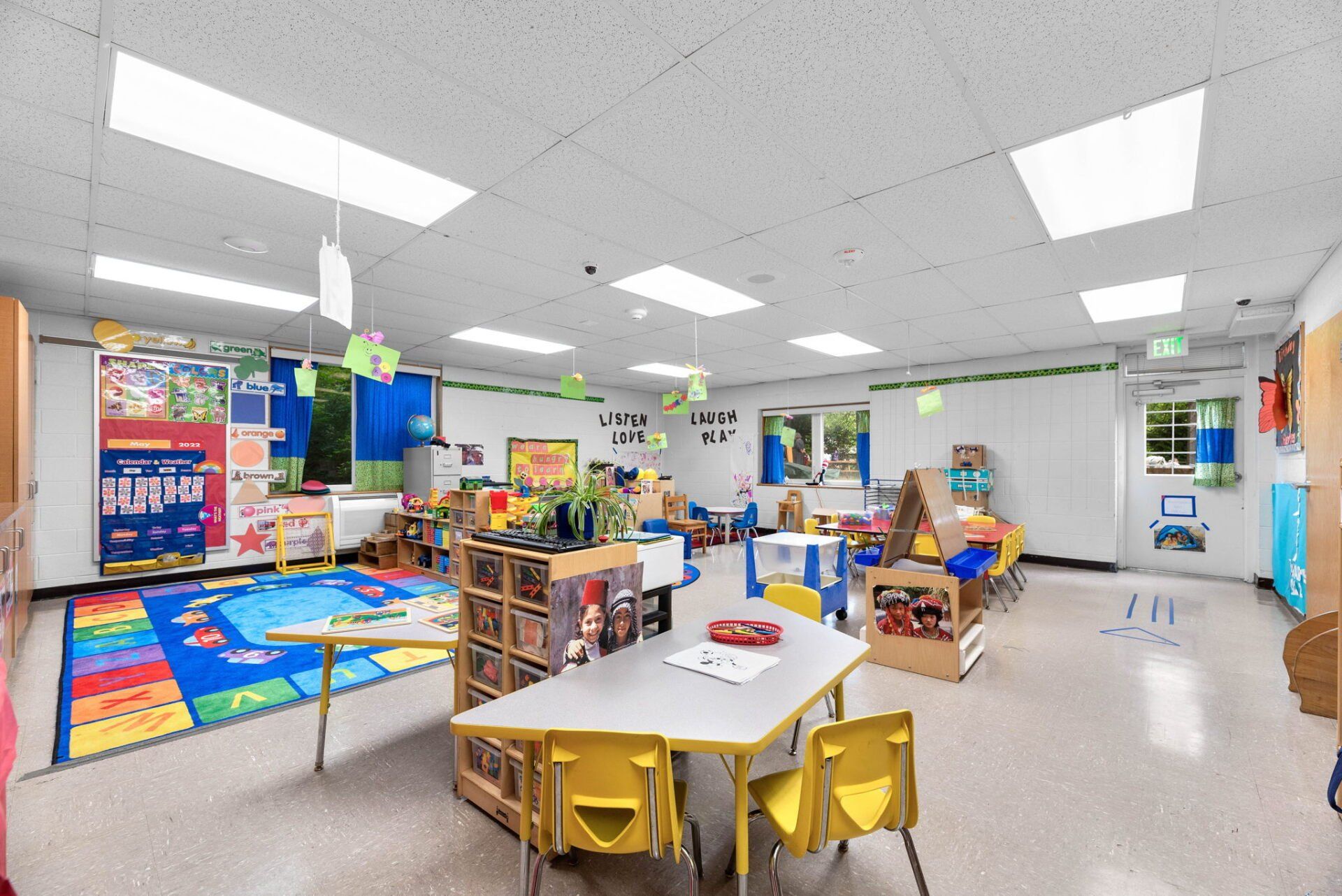 A brightly lit preschool classroom with colorful rugs, tables, toys, and chairs. The space is designed for young children.