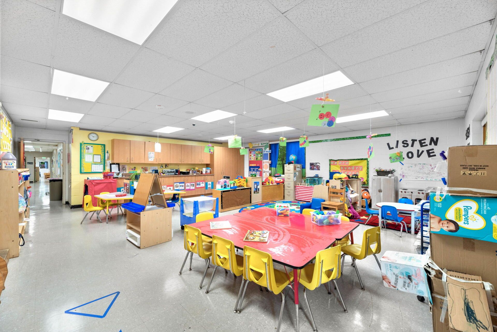 Kindergarten classroom with yellow tables and chairs, art supplies, and decorations; boxes and supplies are stacked in the corners.
