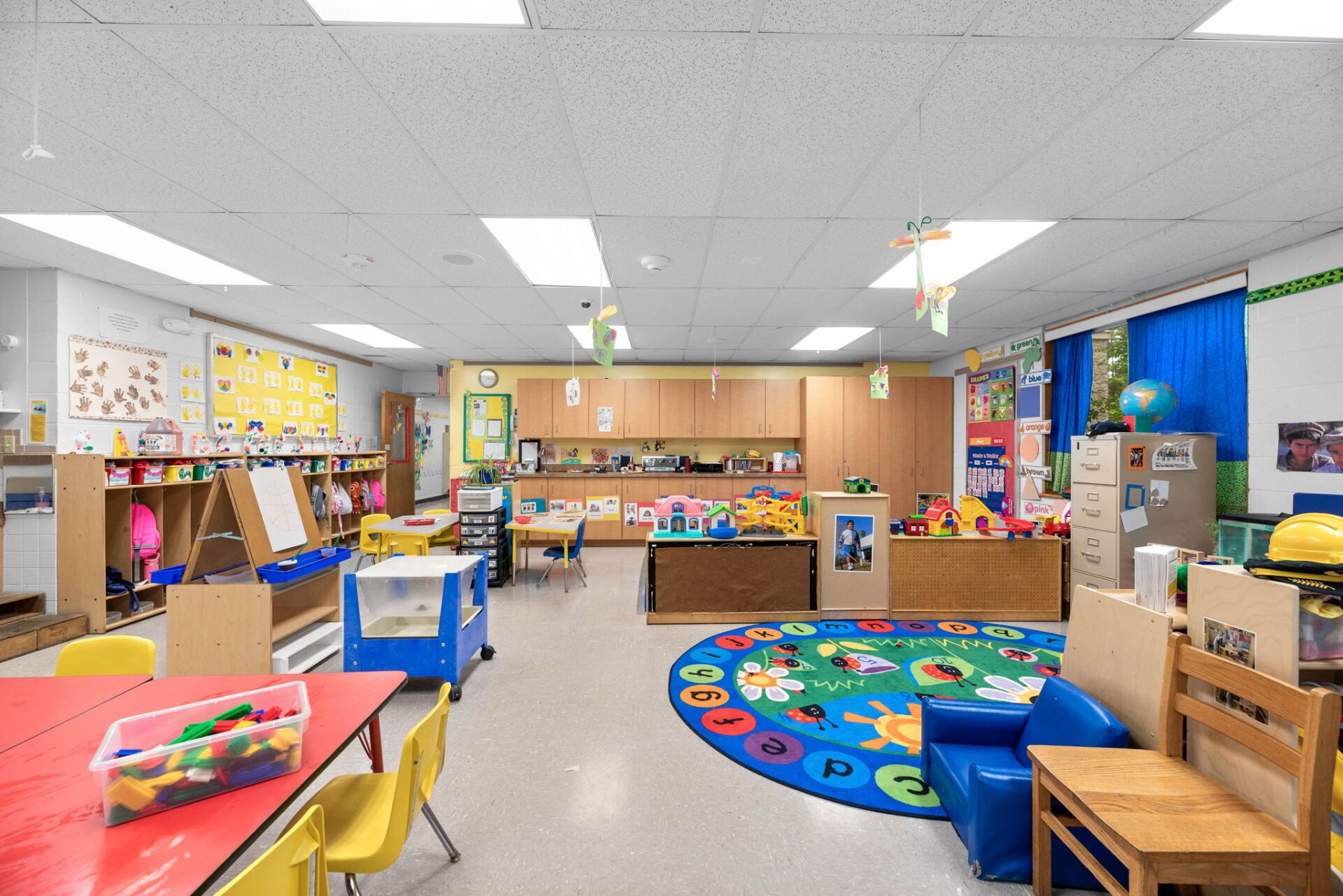 A colorful preschool classroom with tables, toys, bookshelves, and a round rug.