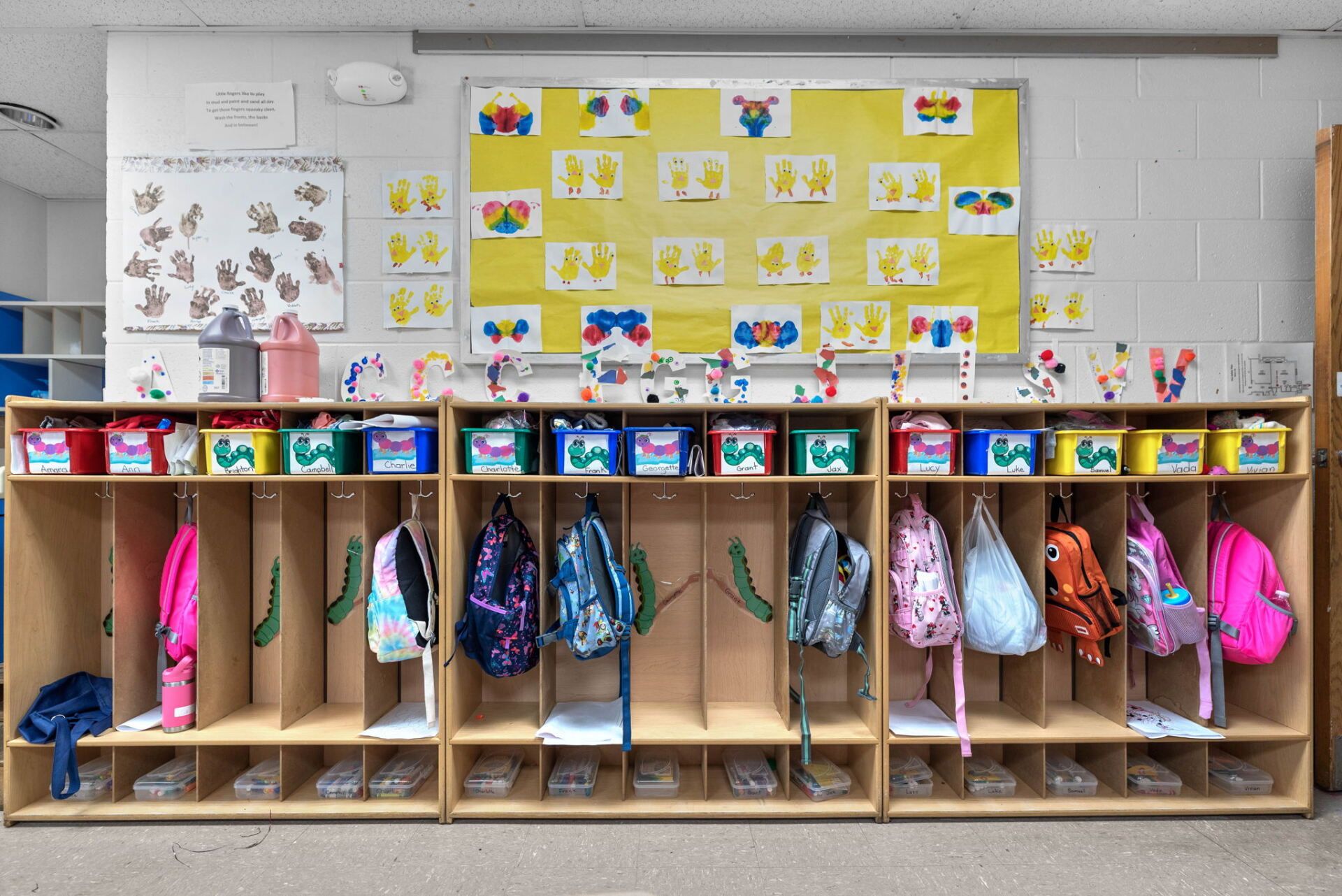 A child's classroom with wooden cubbies, each with a coat, backpack, and bin. Colorful artwork adorns the white wall above.