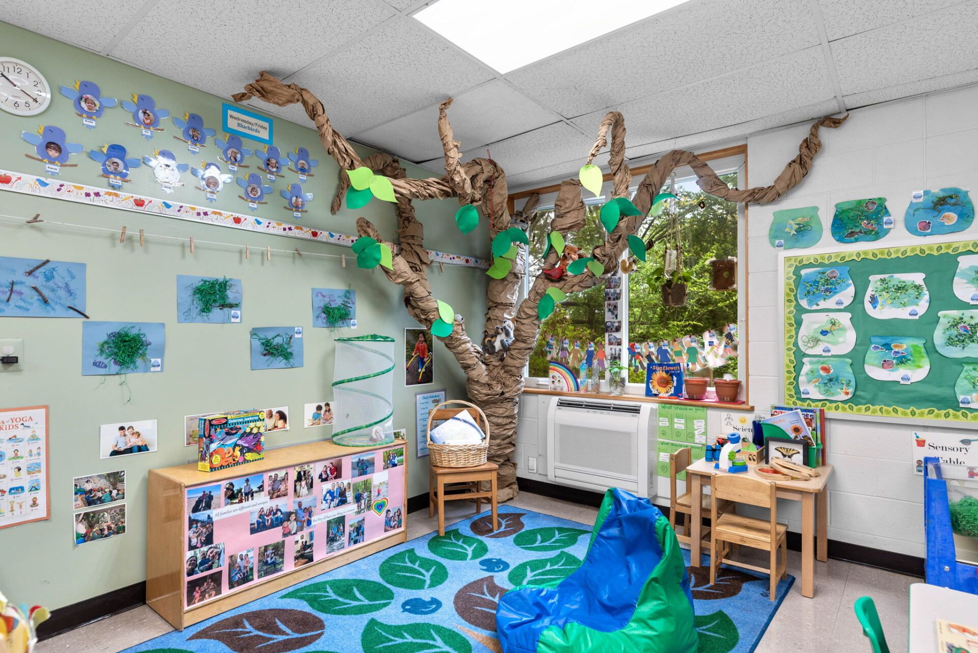 A classroom decorated with a tree made of cardboard. The room has colorful artwork, a rug, and small furniture.
