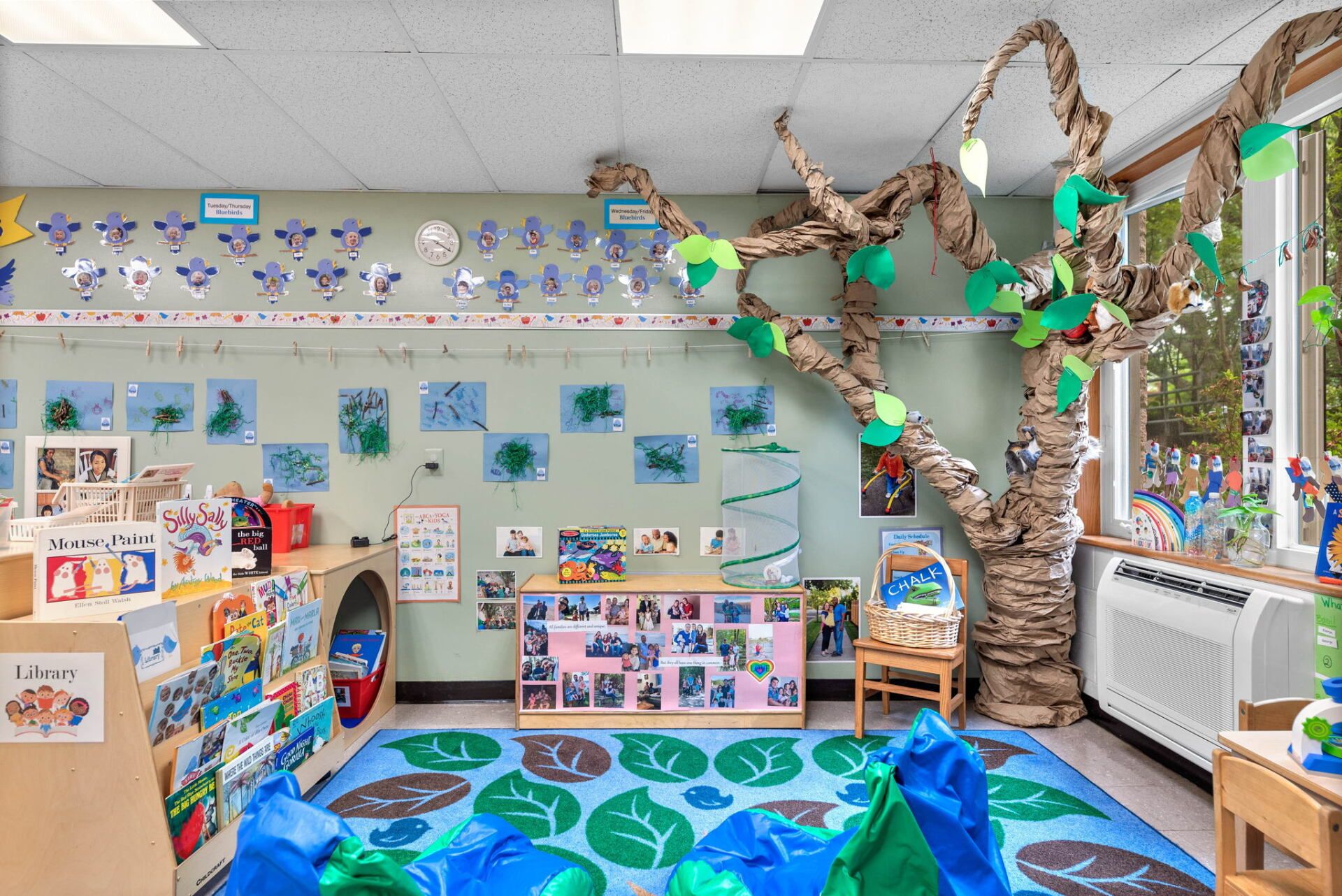 A colorful preschool classroom with a tree-like structure, leaf-shaped rug, and student artwork on the walls.