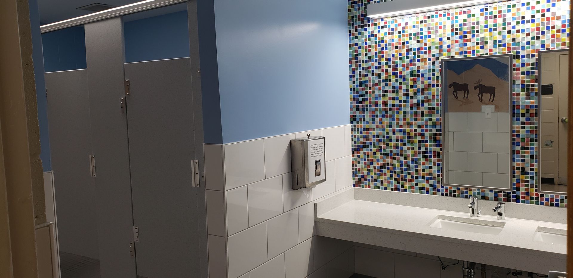 Bathroom interior featuring stalls, a sink, mirror, and colorful tile wall. Light blue and white colors dominate.