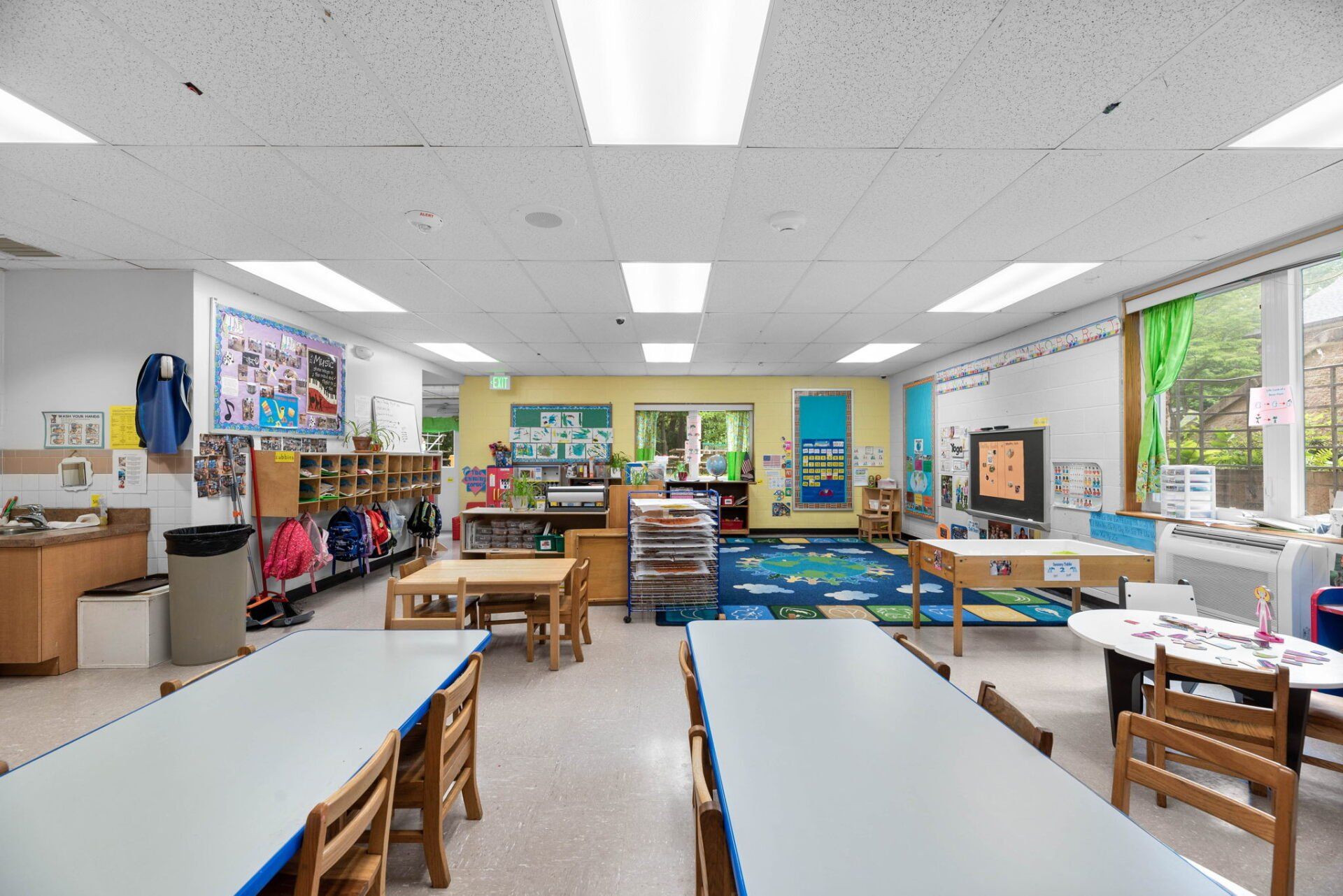 A bright, colorful preschool classroom with tables and chairs. Displays and windows are visible.