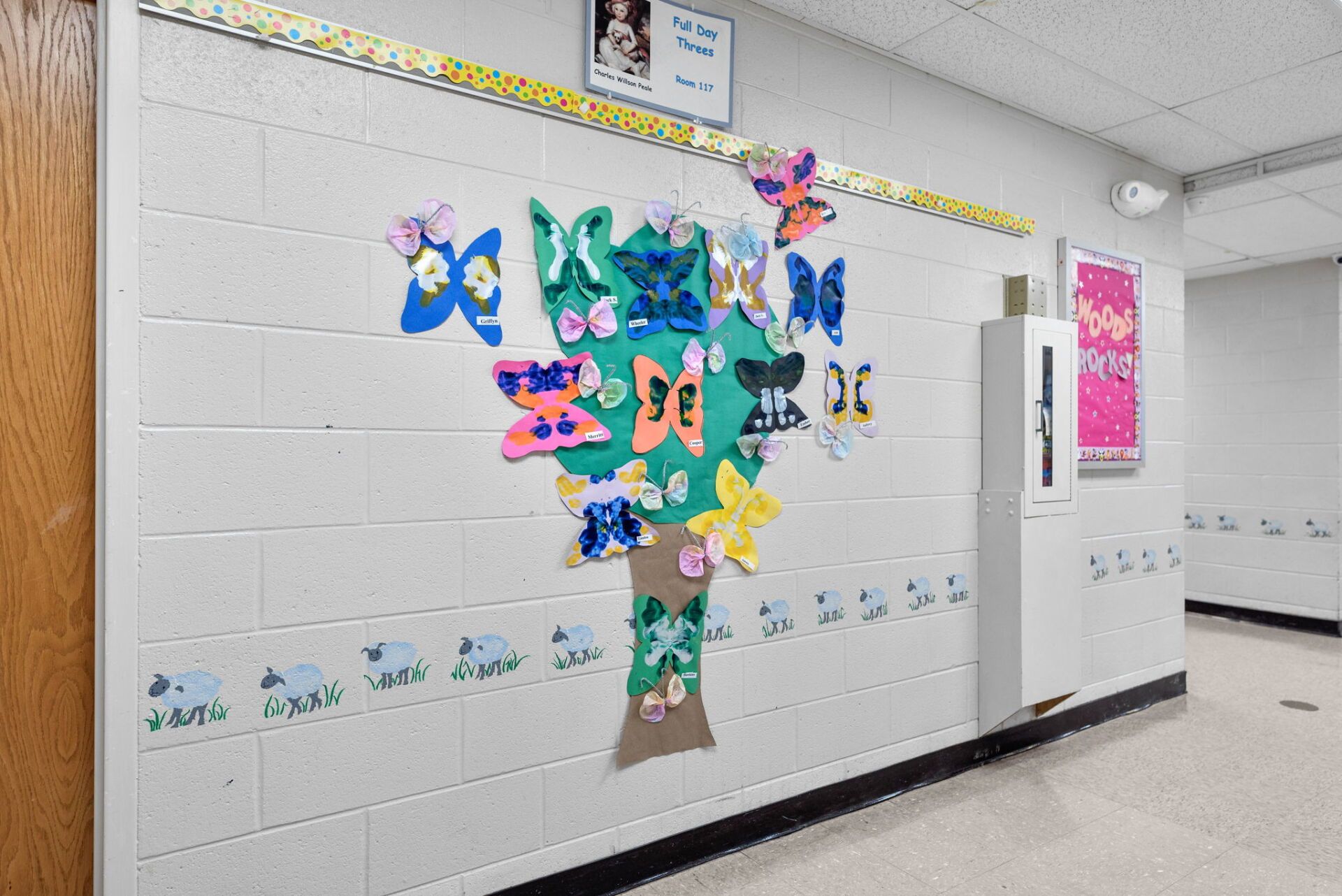 A classroom hallway with a decorated tree of butterflies.  Butterflies of various colors are attached to a brown paper tree on a white brick wall.