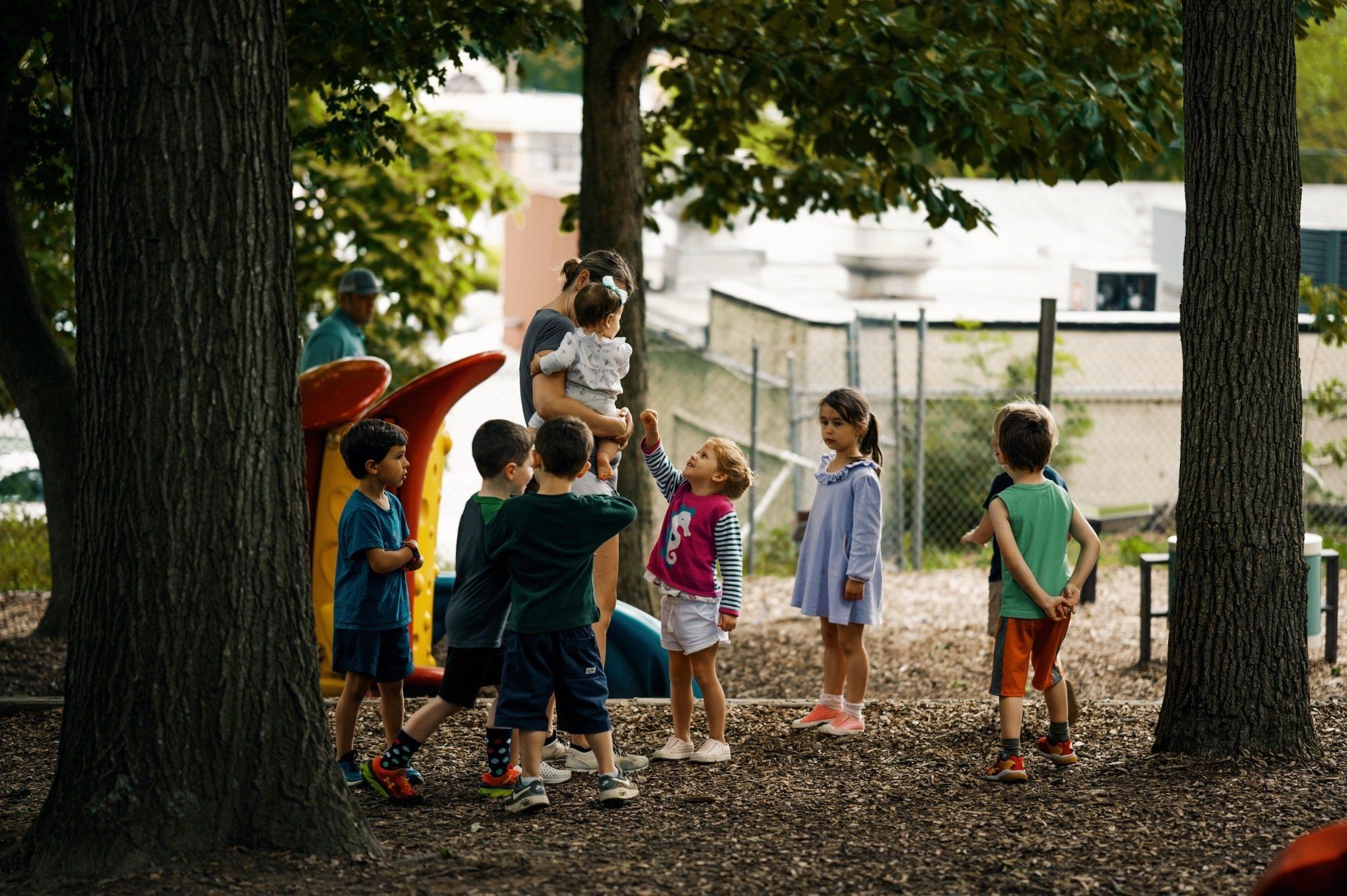 Children gather around a caregiver, reaching up in a park setting with trees, a playground, and a building in the background.