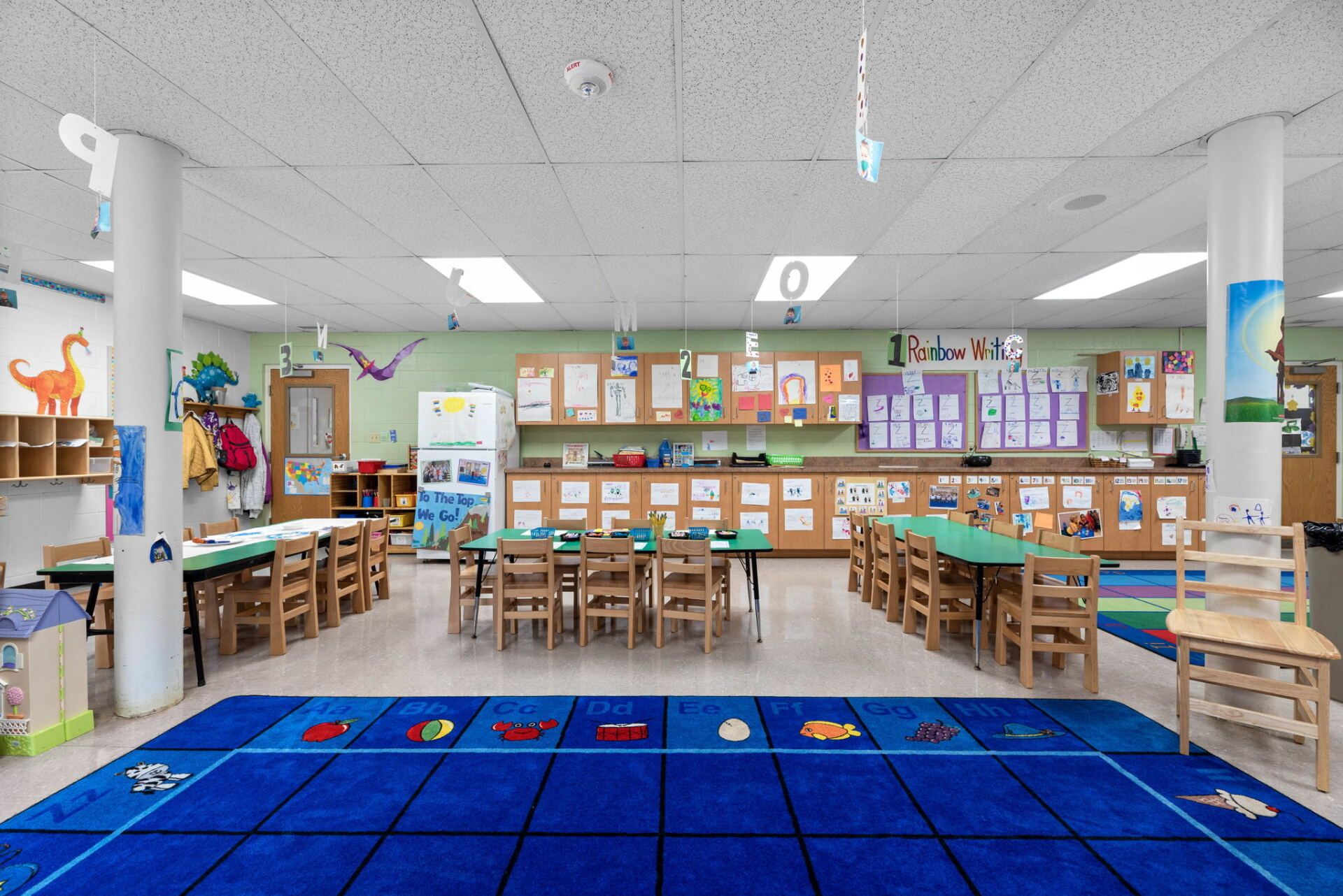 A preschool classroom with tables, chairs, and learning materials. Blue rug on the floor, white pillars.