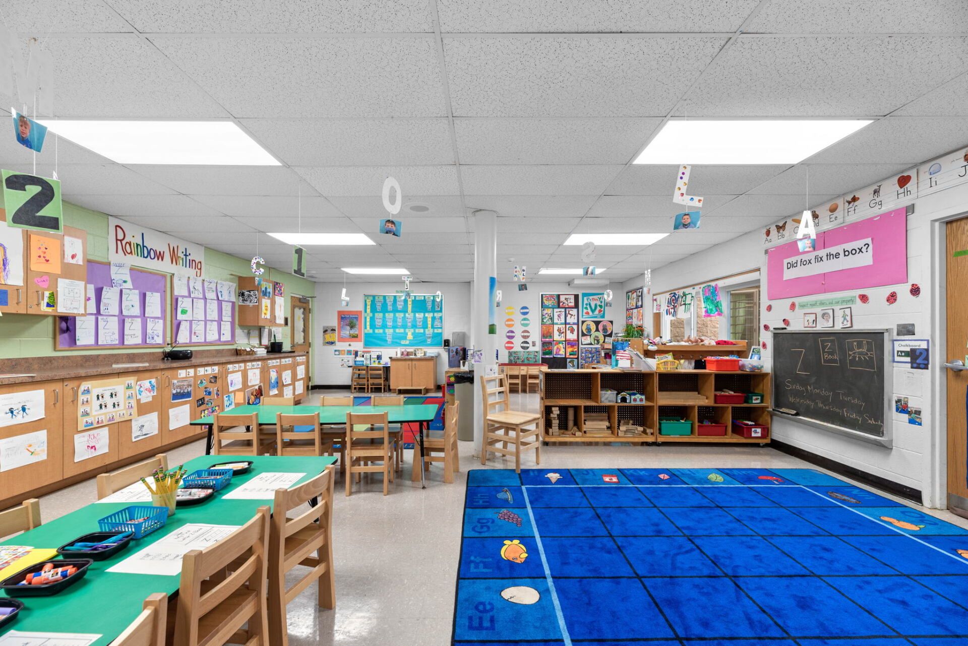 A brightly lit kindergarten classroom with small tables, chairs, and a blue rug. Artwork and learning materials decorate the walls.