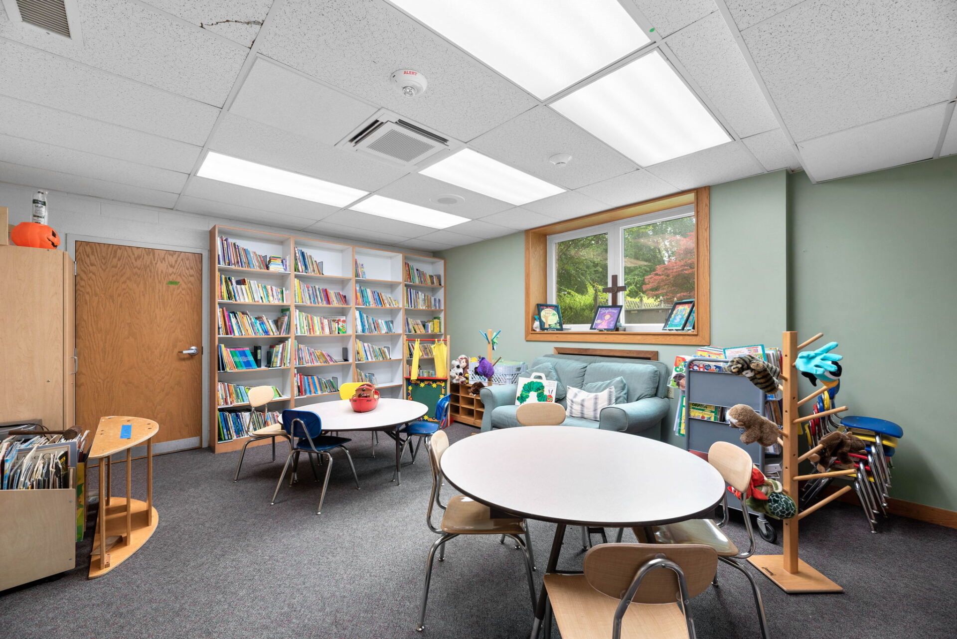 A brightly lit children's reading room with bookshelves, small tables and chairs, a sofa, and a window looking outside.