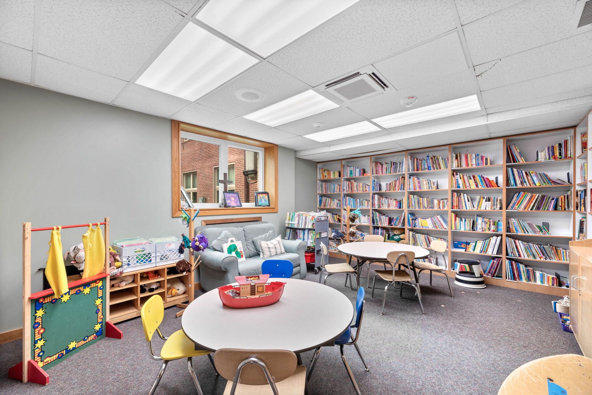 A brightly lit children's library with bookshelves lining the walls, round tables, small chairs, and a reading nook.