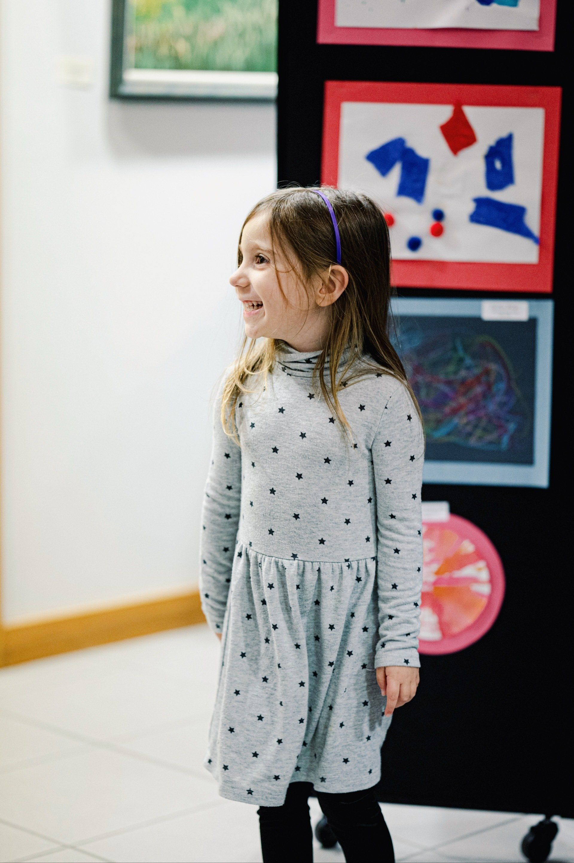 Smiling young girl in a gray dress with black leggings, looking to the side near artwork on a black display.