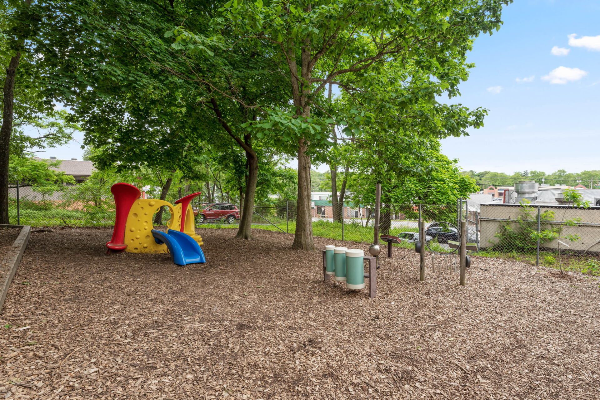 Playground with a colorful slide, musical instruments, and trees, set on wood chips under a partly cloudy sky.