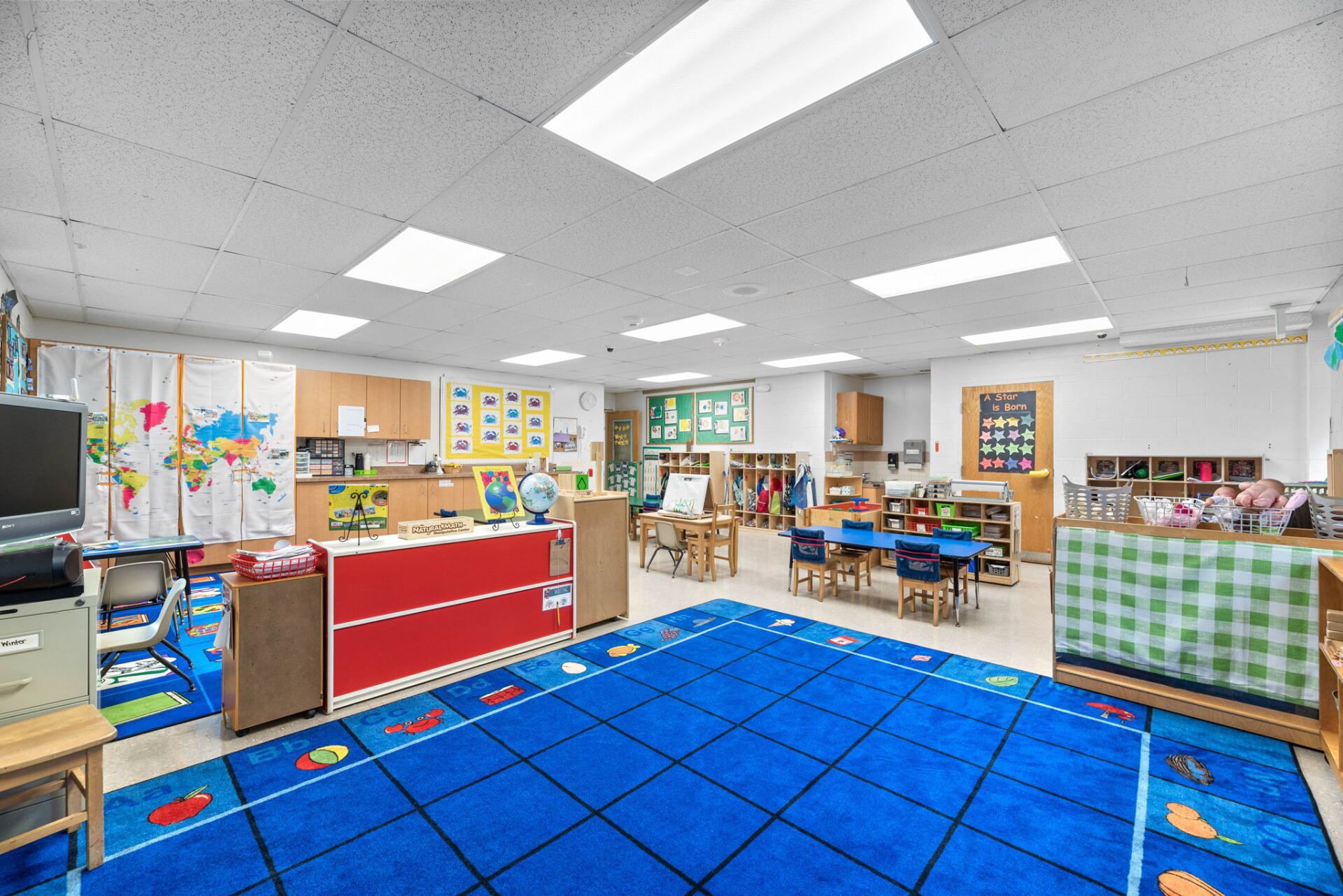 A brightly lit preschool classroom with a blue rug, red cabinets, and tables and chairs for children.