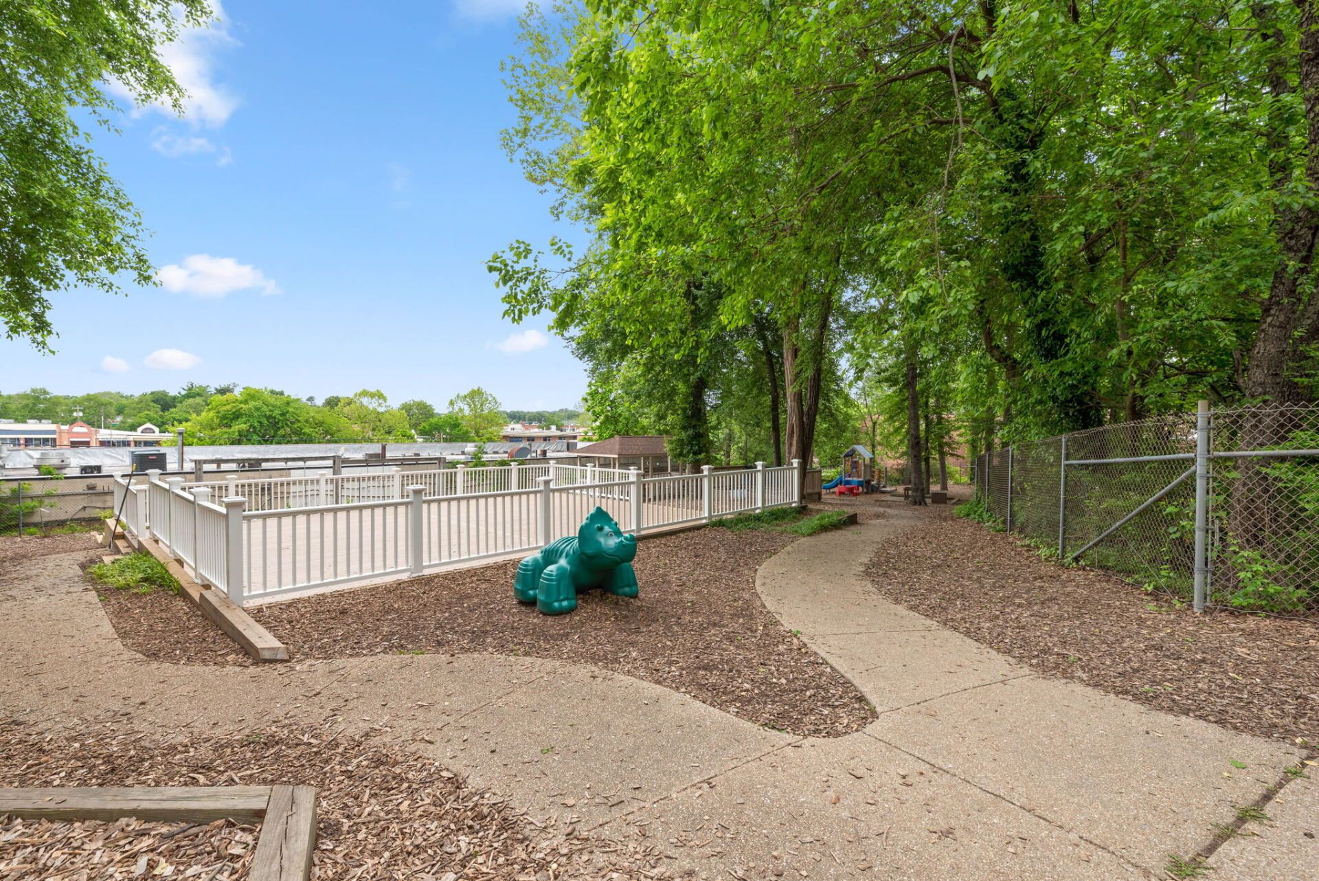 A dog park with a paved path, white fenced area, green play structure, and surrounding trees.
