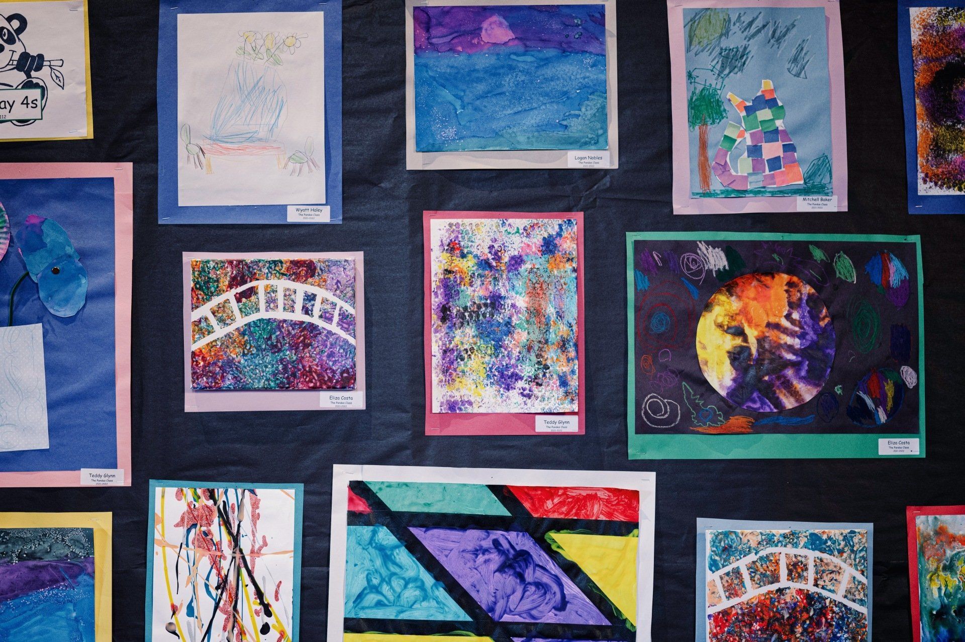 Artwork display featuring colorful drawings, including bridges, landscapes, and abstract pieces, each framed with colored paper.