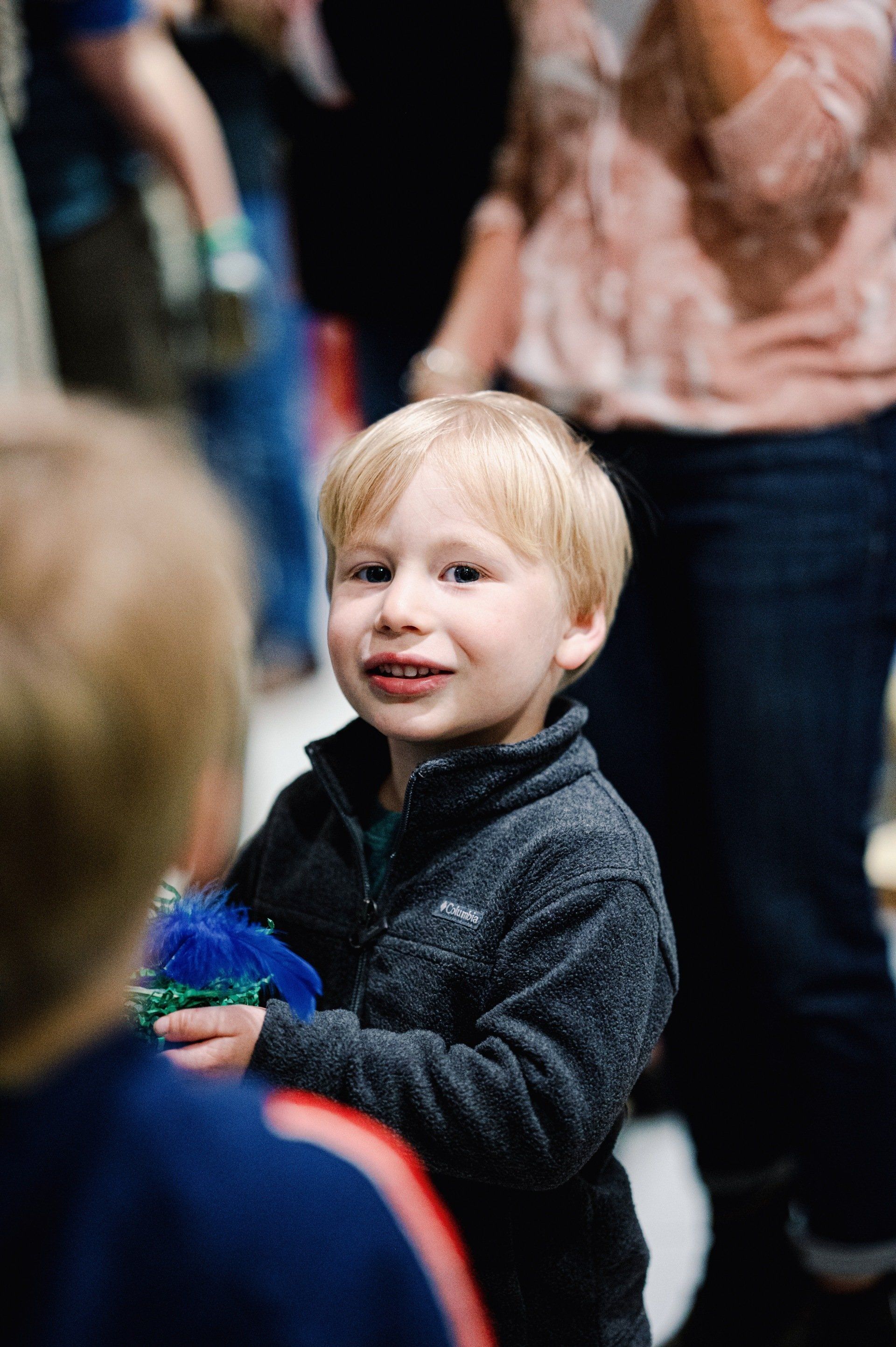 A young boy with blonde hair smiles, wearing a dark jacket, holding a toy, and looking towards the camera; other people are in the background.