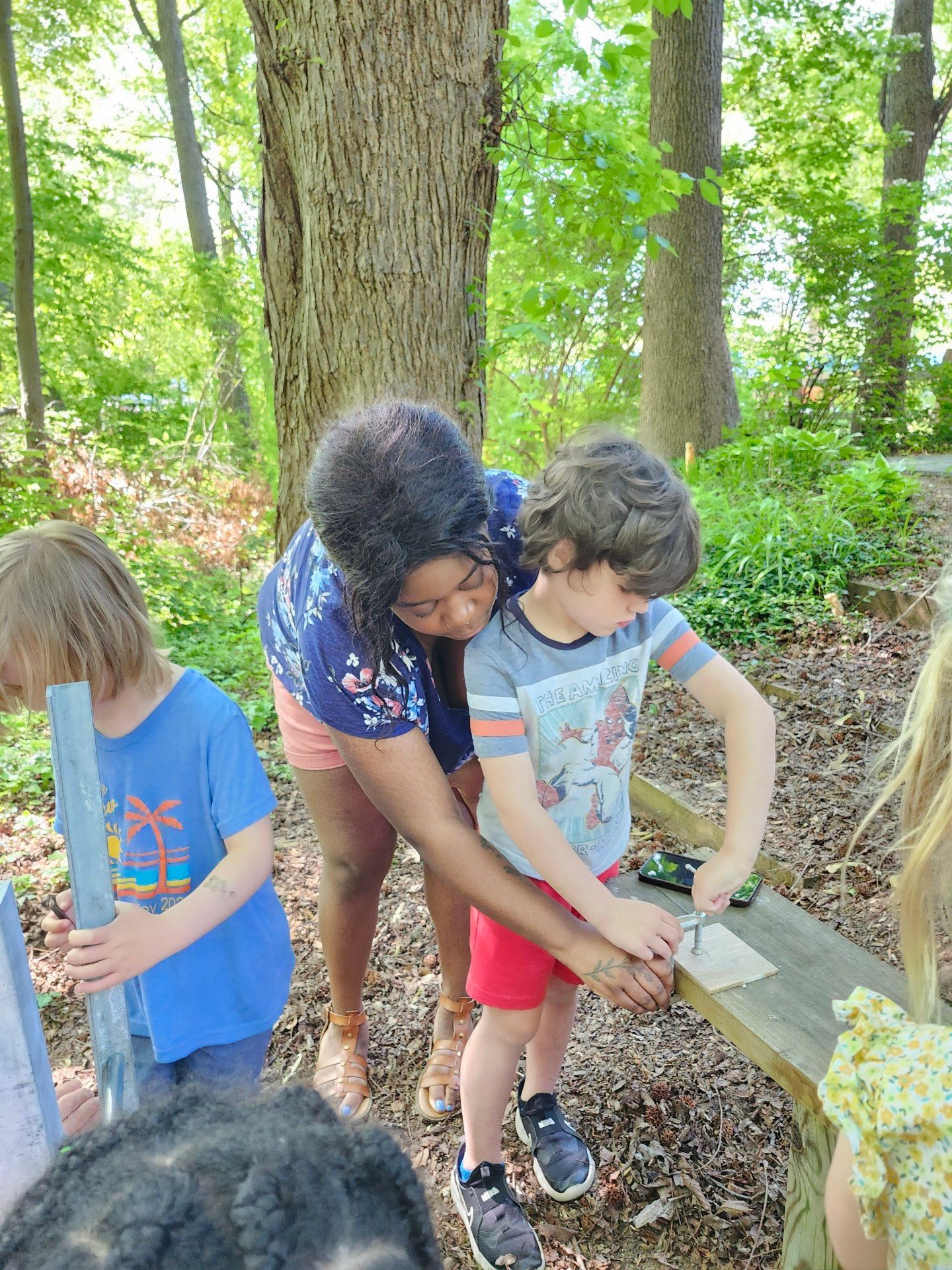 A woman helps a young boy with a small tool at a wooden bench outdoors, other children watch.