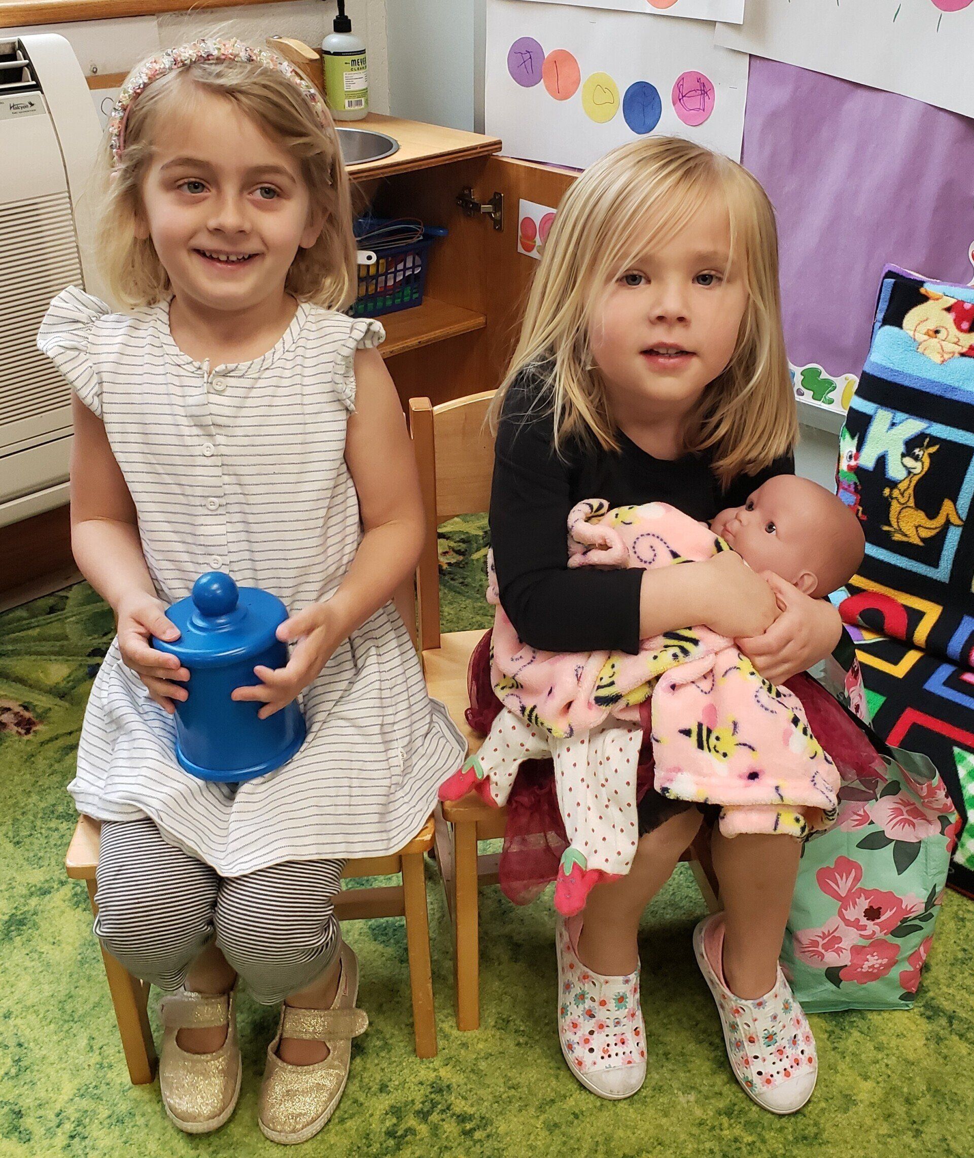Two young girls sit on small chairs, one holding a blue jar, the other cuddling a doll. They are in a colorful playroom.
