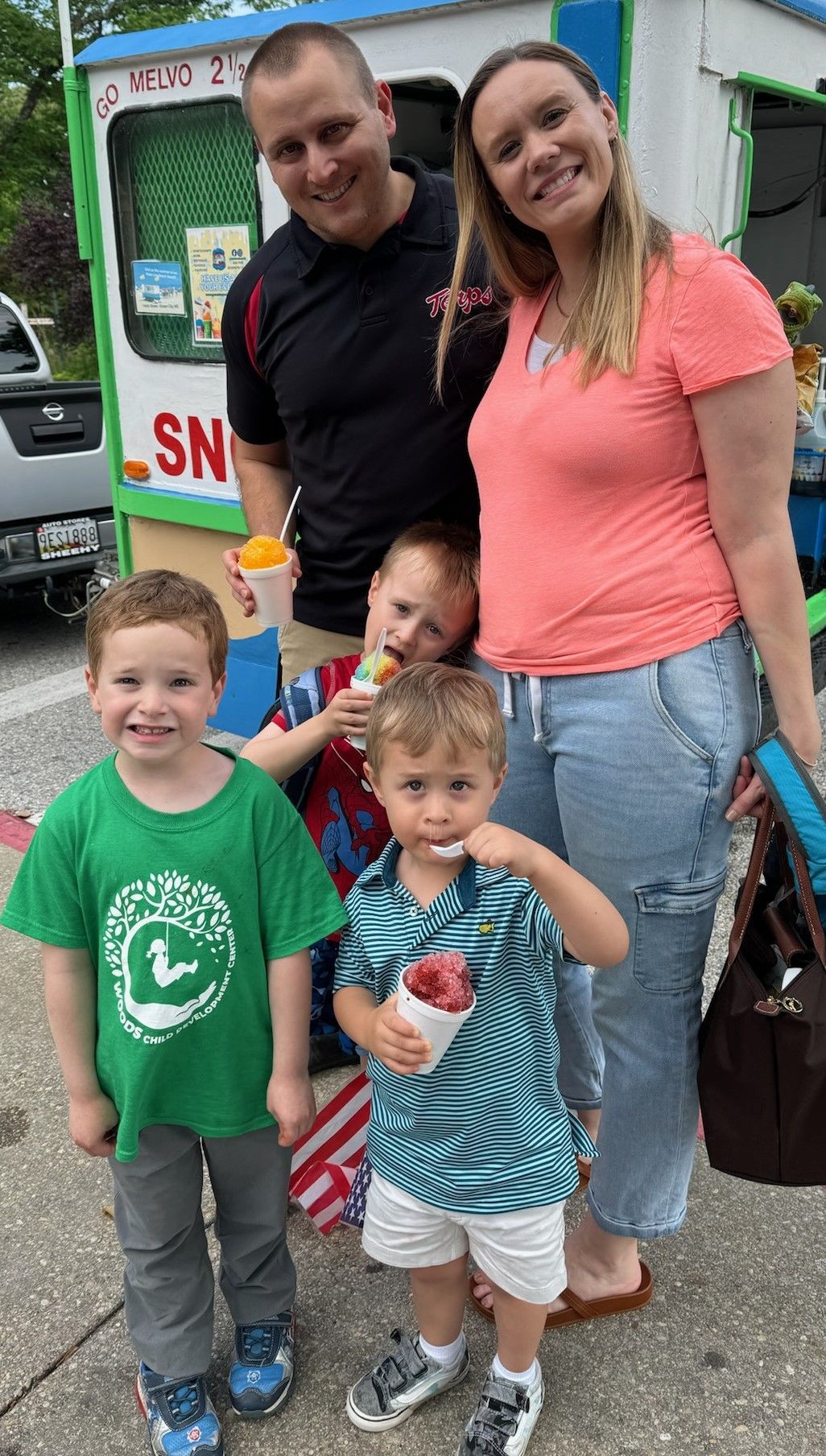 Family of five smiles at a snow cone stand: Two parents, three young children, enjoying treats on a sunny day.