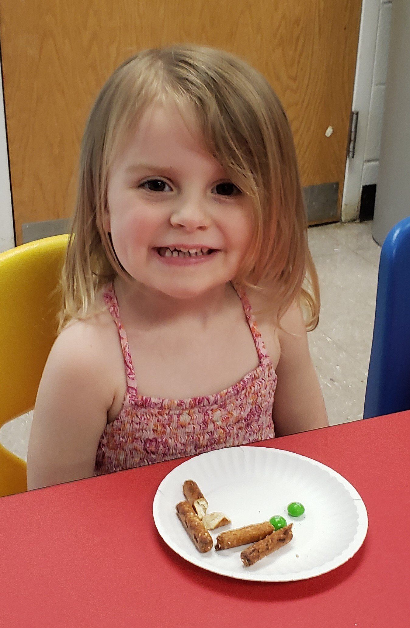 A young girl with blonde hair and a pink patterned top smiles at the camera, a plate of snacks on the red table in front of her.