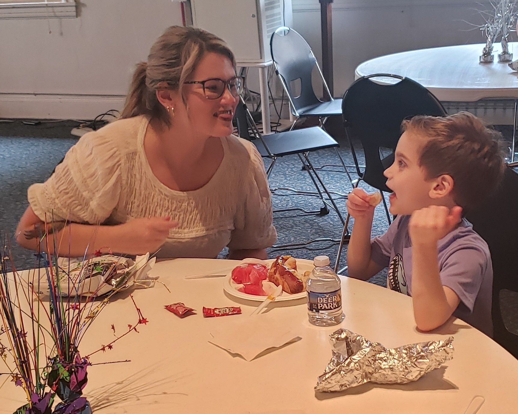 Woman with glasses smiles at a young boy who is excitedly talking at a round table. They are at a party, with food and decorations.