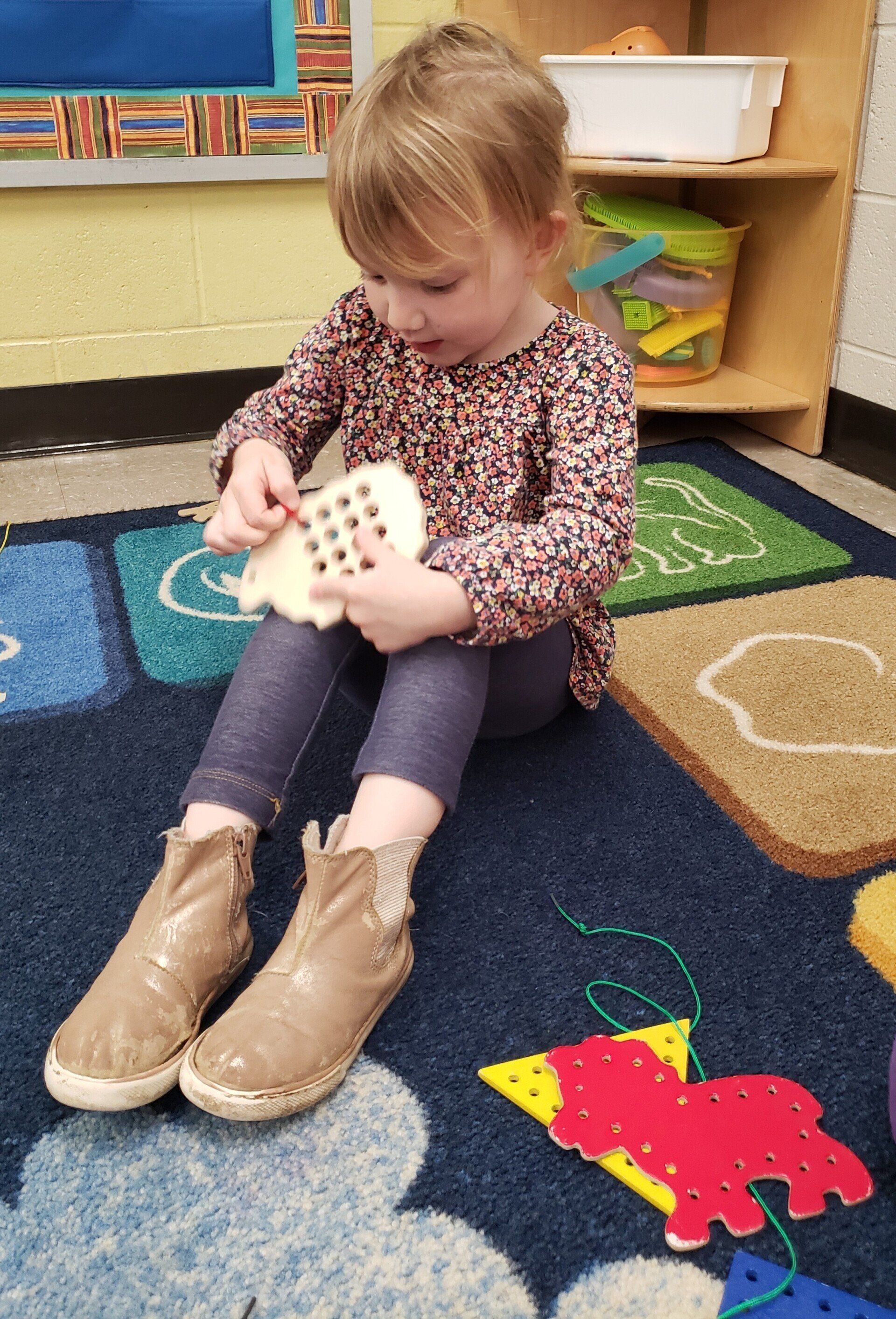 A young child sits on a rug and focuses on lacing a toy shaped like a bear. She wears a patterned top and boots.