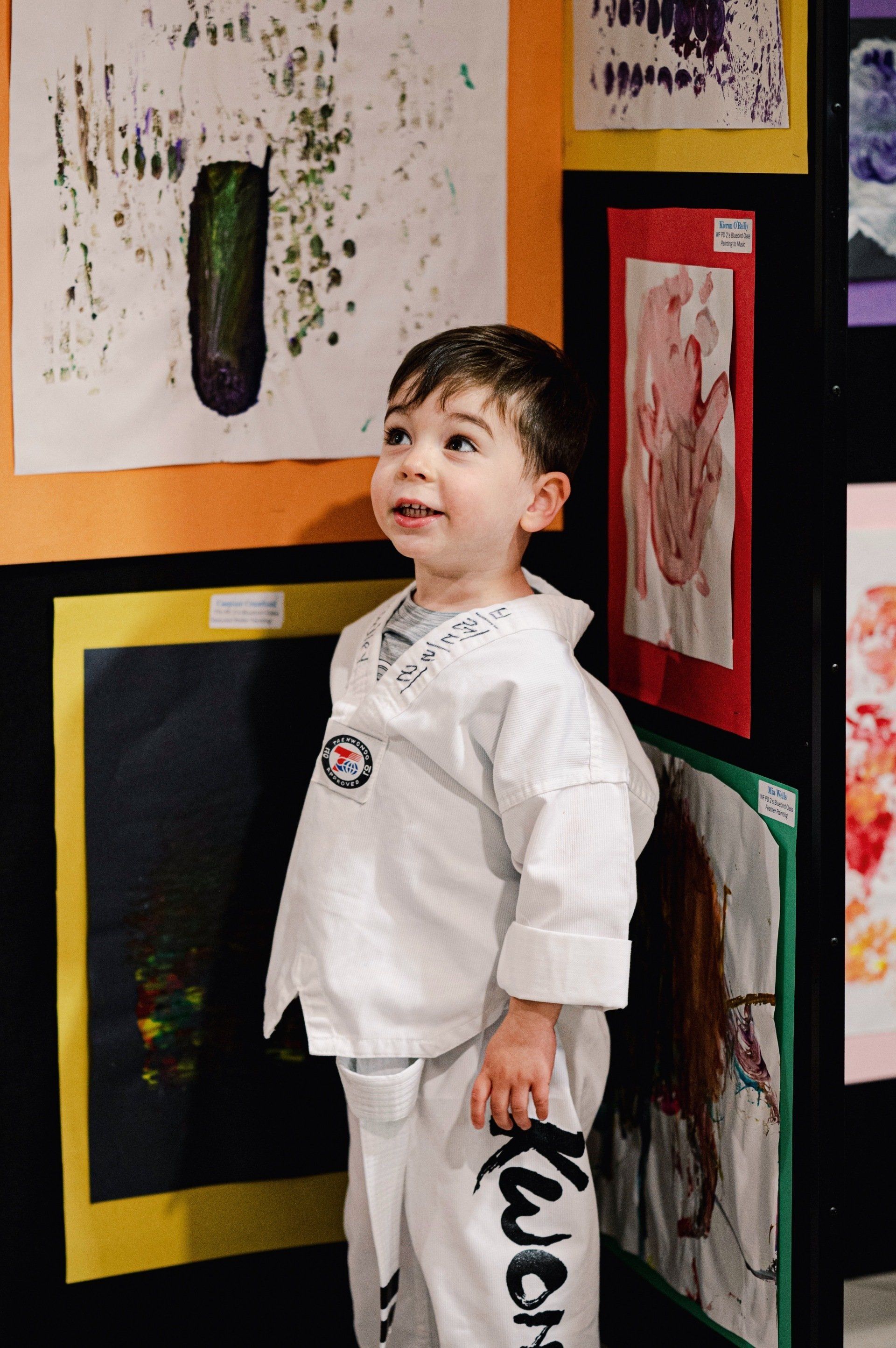 A young boy in a taekwondo uniform admires artwork at an exhibition, smiling and looking upward.
