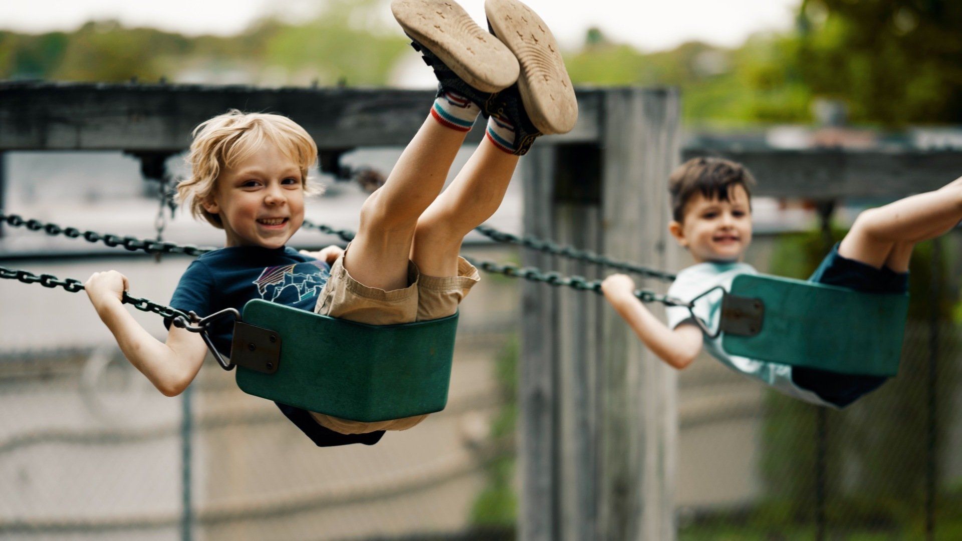 Two young boys swinging high at a playground. Smiling, one has blonde hair, other has dark.