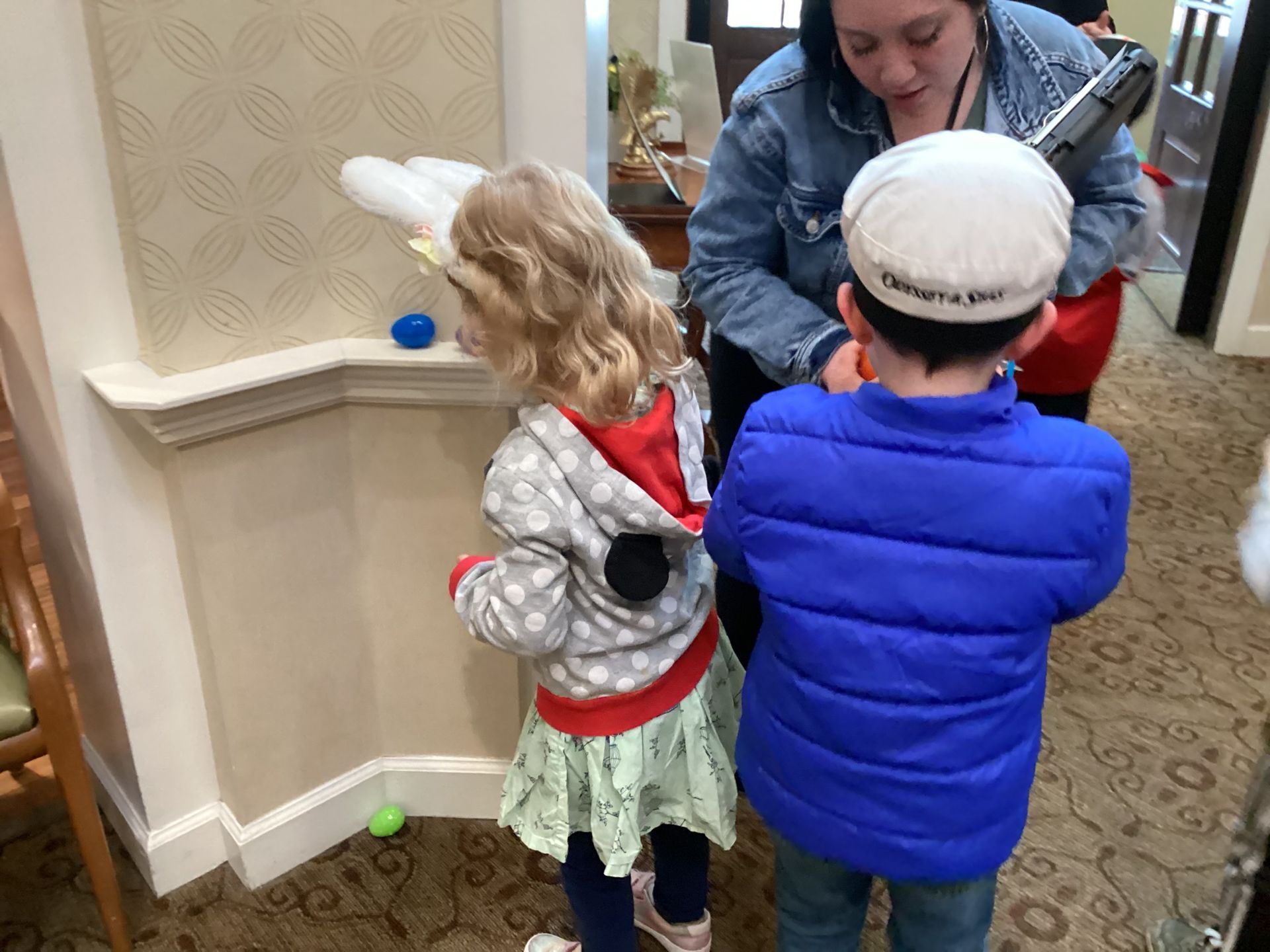 Two children searching for Easter eggs, guided by a woman, indoors near a wall with decorations.