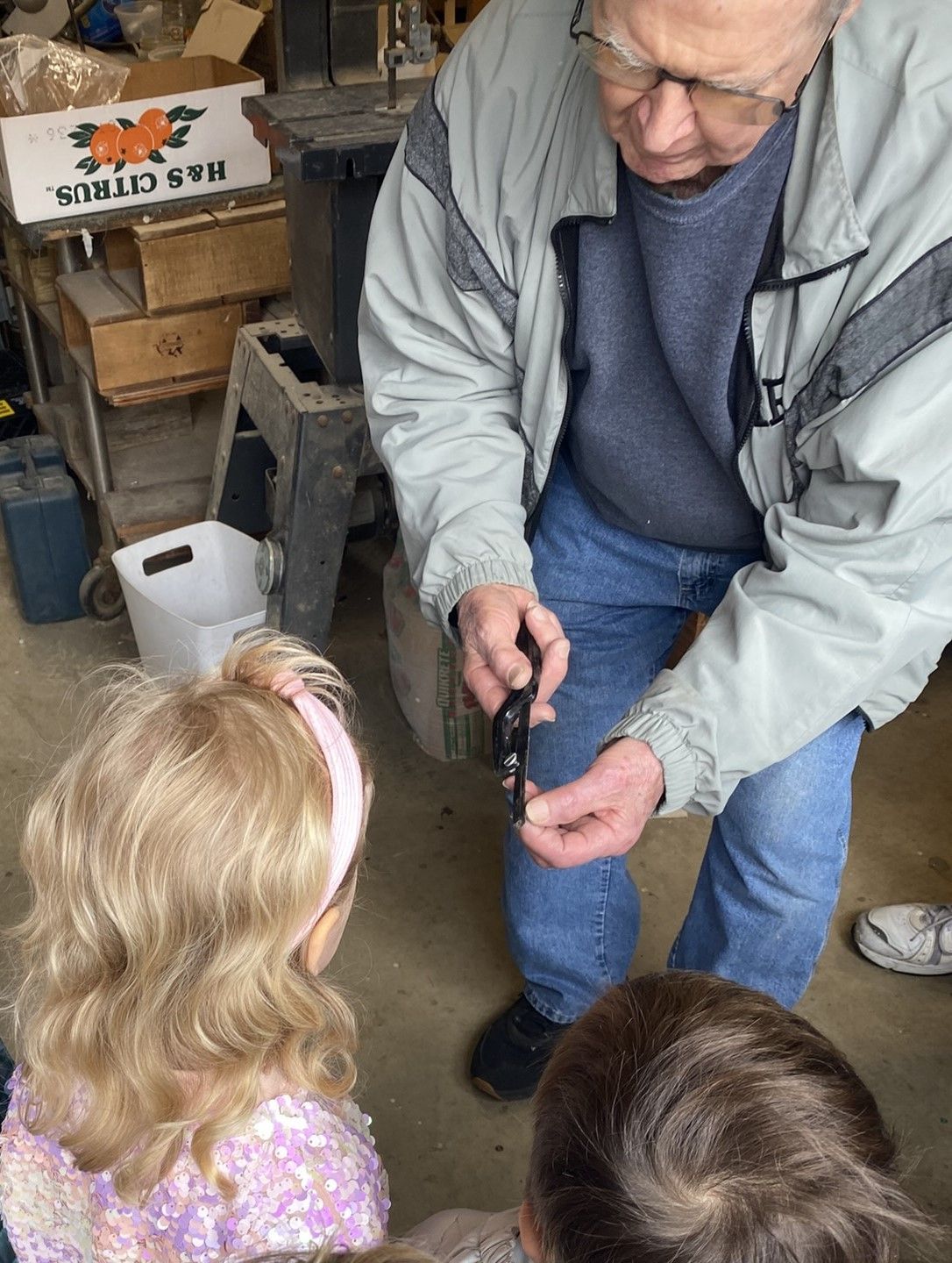 An elderly man shows two children a small object. They are indoors, likely in a garage or workshop.