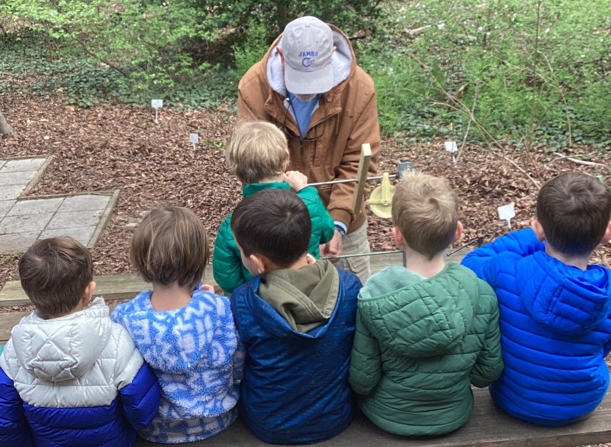 A man shows a brass instrument to a line of children in a wooded outdoor setting. The children are wearing colorful jackets.