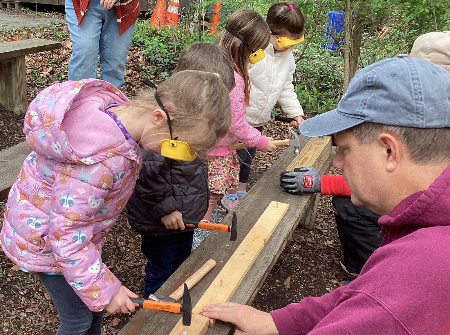 Children hammering nails into wood with an instructor. They are wearing eye and ear protection outdoors, in a forest setting.