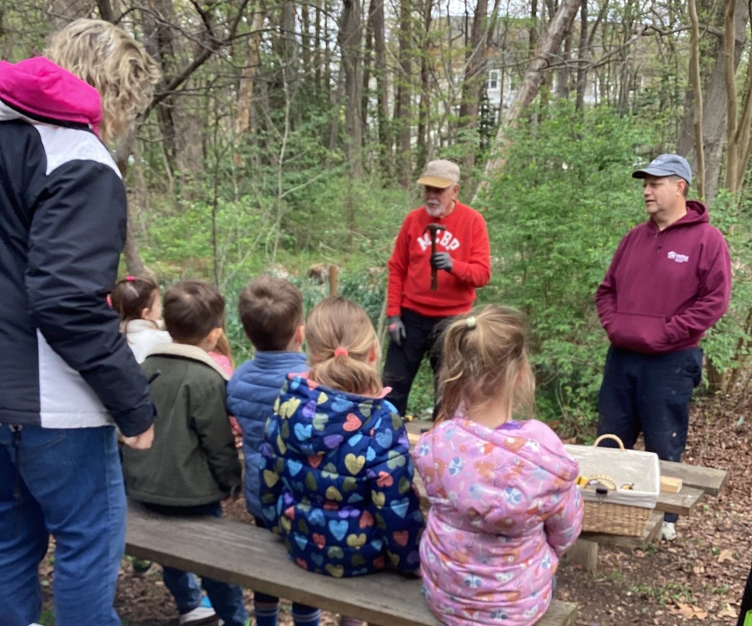 A group of young children and adults listen to a man in a red sweatshirt in an outdoor, wooded setting.