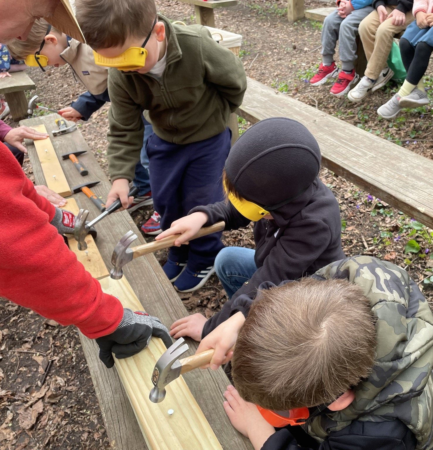Children hammering nails into wood planks outdoors. They wear safety glasses and are supervised by adults.