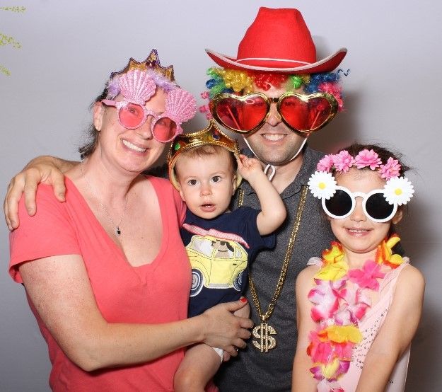 Family in fun photo booth with silly glasses and hats; two adults, two kids, smiling.