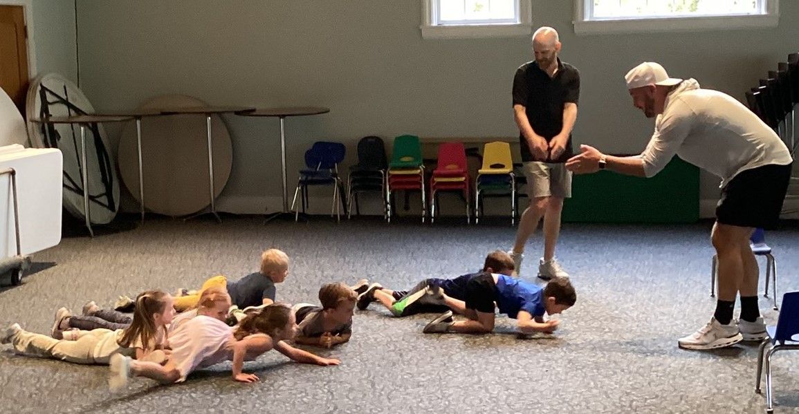Children crawling on the floor with two adults supervising in a room with chairs and tables.