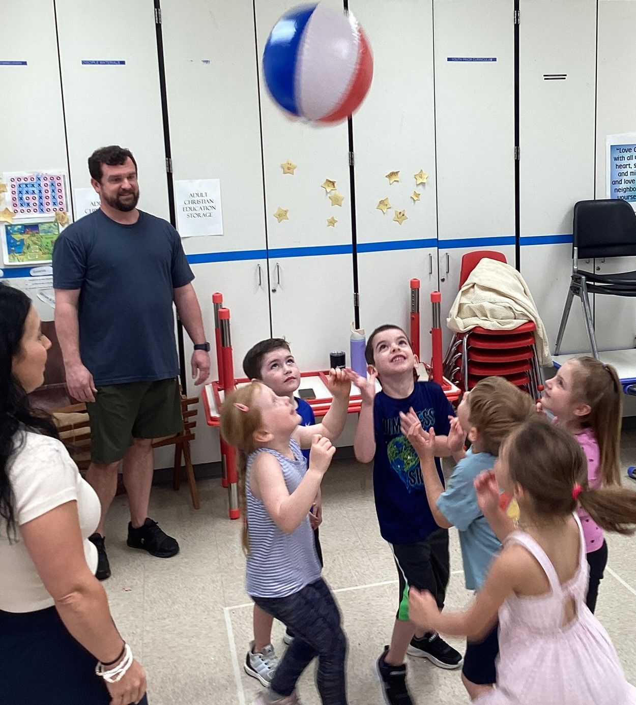 Children play with a beach ball in a classroom, watched by two adults. The ball is blue, white, and red.