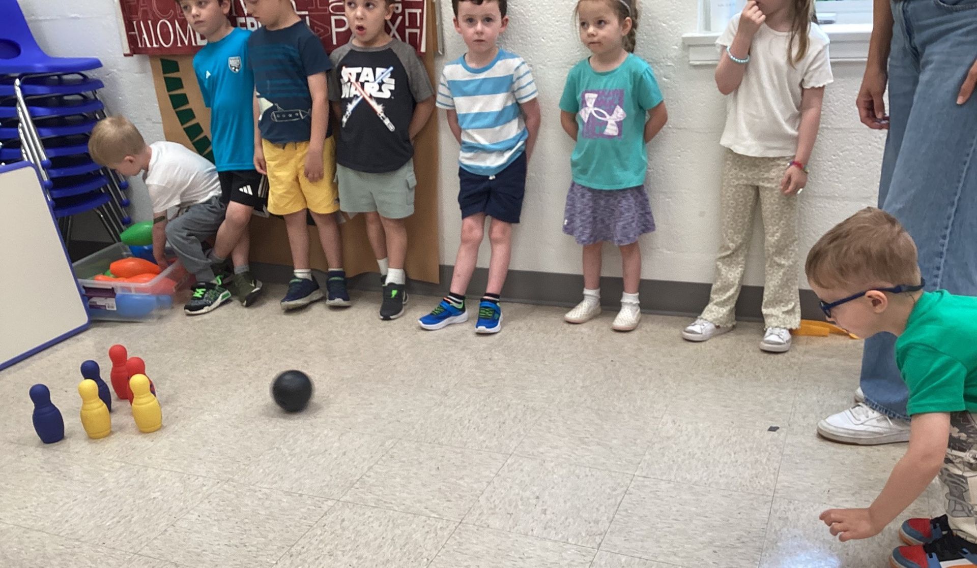 Children playing mini-bowling indoors. A boy rolls a black ball toward colorful pins while others watch.