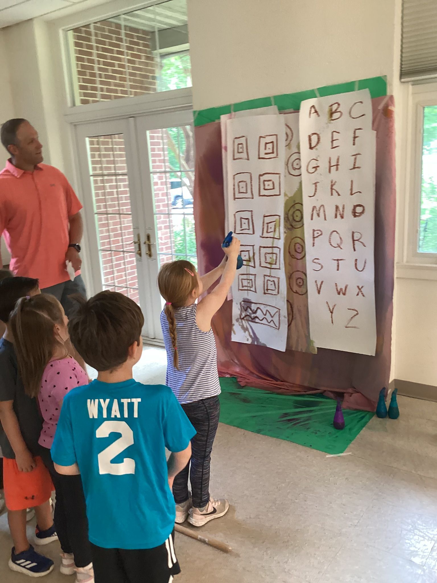 A group of young children spray paint stencils on paper with guidance from an adult in a classroom setting.