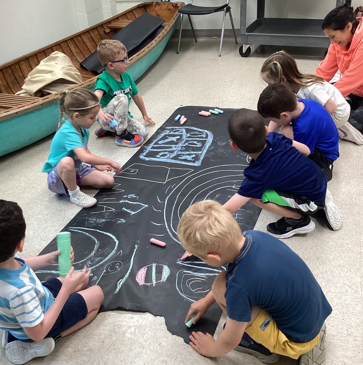 Children drawing with chalk on a large, dark sheet on a floor. A canoe and seated adult are nearby.