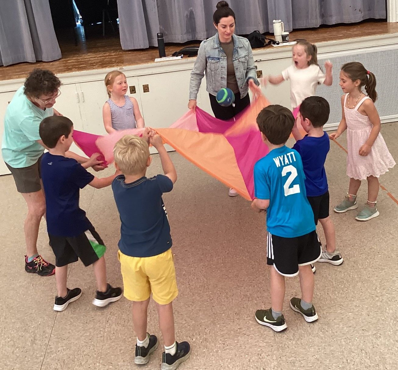 Children and adults playing with a colorful parachute indoors. They are holding the edges and bouncing a ball on it.