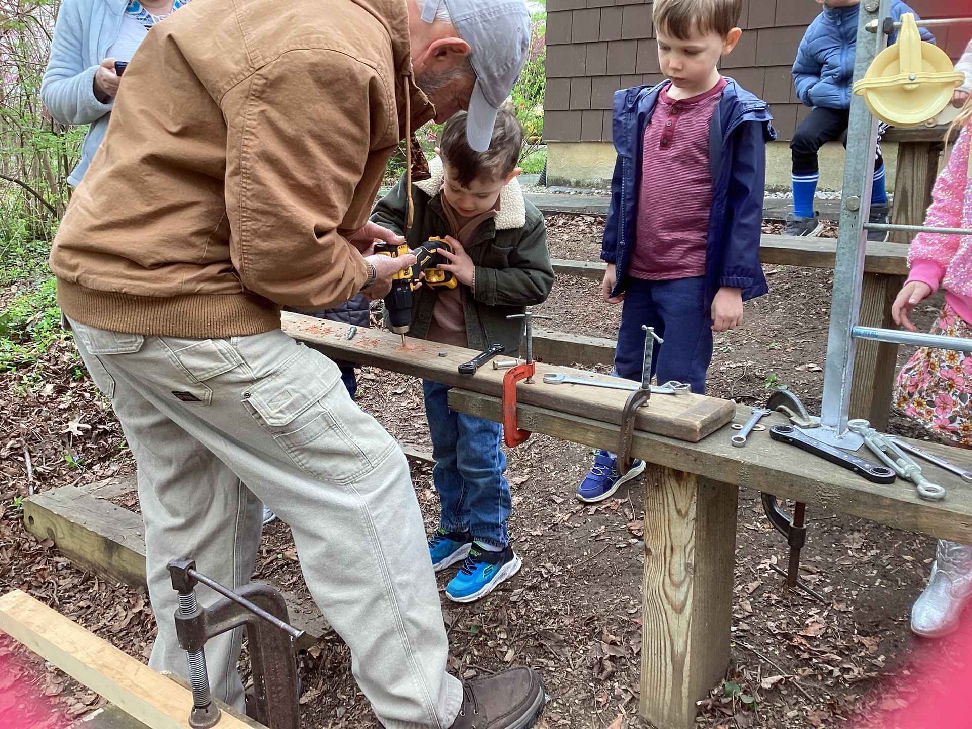 A man shows two young boys how to work on a piece of wood outdoors. The boys watch intently.